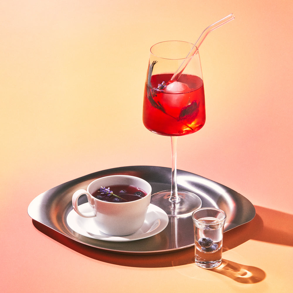 OSULLOC Hibiscus Sunshine — Red iced infusion in a stemmed glass and a cup of dark infusion on a metal tray.