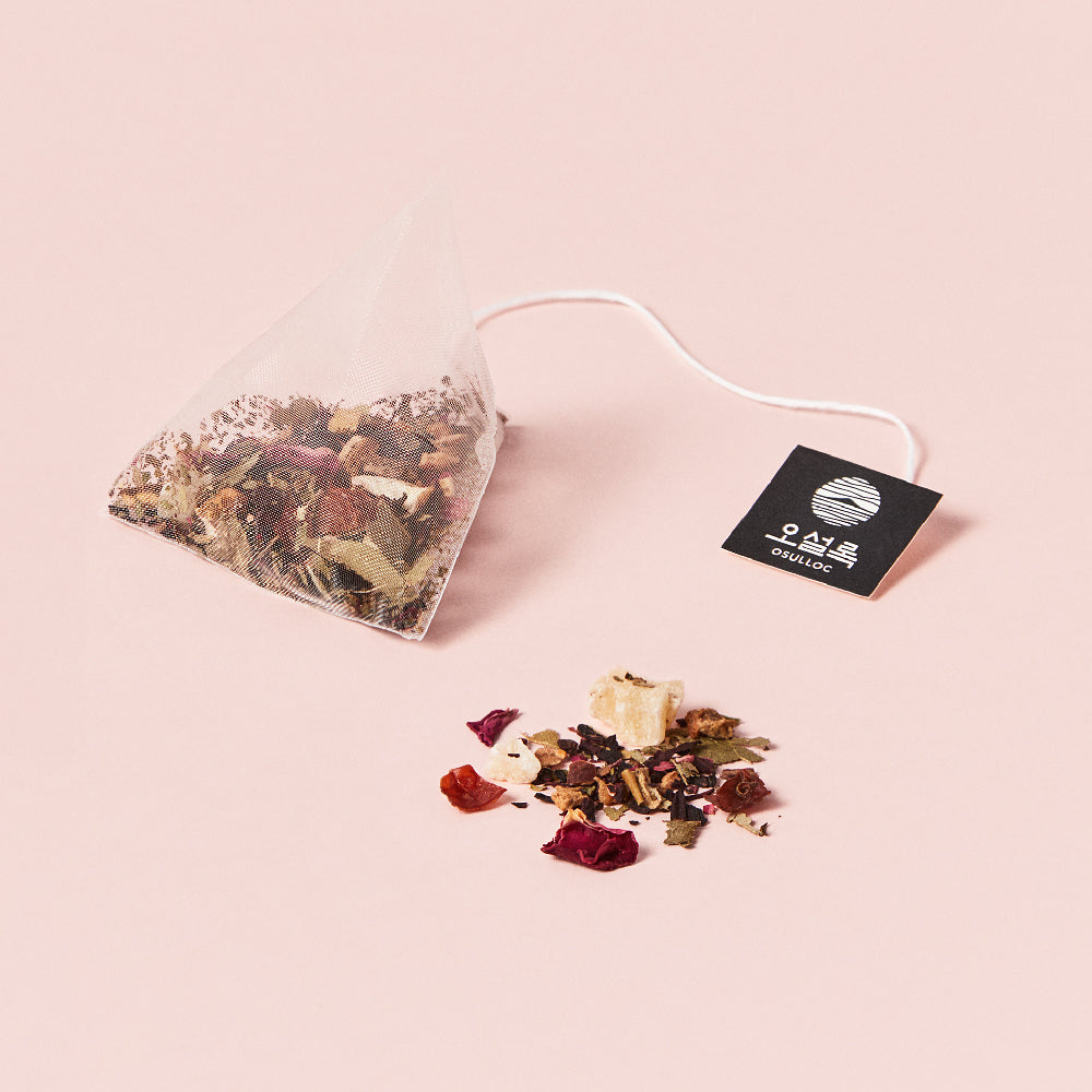 OSULLOC Hibiscus Sunshine — Pyramid tea bag and scattered fruit-and-flower blend on pink.