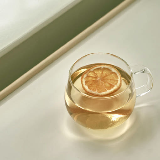 Glass cup of pale golden tea with a dried citrus slice on a sunlit windowsill.