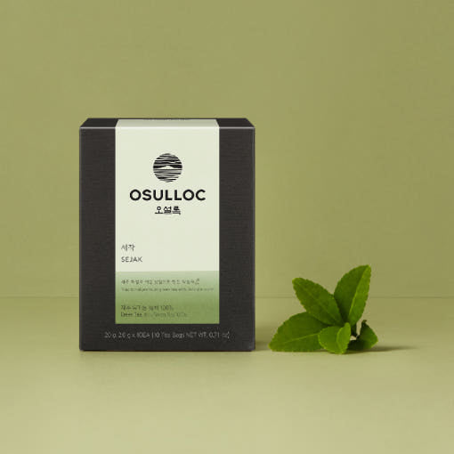 OSULLOC Sejak Green Tea — Dark Sejak box beside fresh green leaves on an olive background.