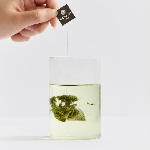 OSULLOC Sejak Green Tea — Hand holding the tag of a tea bag steeping in a glass of pale green tea.