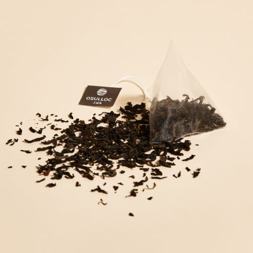 OSULLOC Jeju Volcanic Rock Tea — Bolsita piramidal junto a hojas oscuras de té esparcidas.