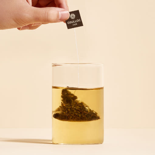 OSULLOC Jeju Volcanic Rock Tea — Mano sujetando la etiqueta de una bolsita piramidal que infusiona en té dorado.