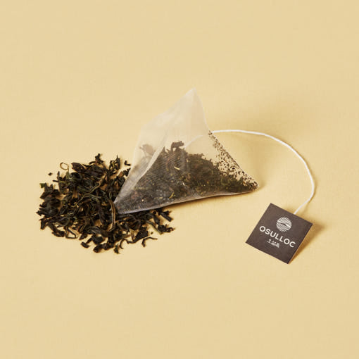 OSULLOC Green Oolong — Pyramid tea bag beside loose dark leaves on ochre.