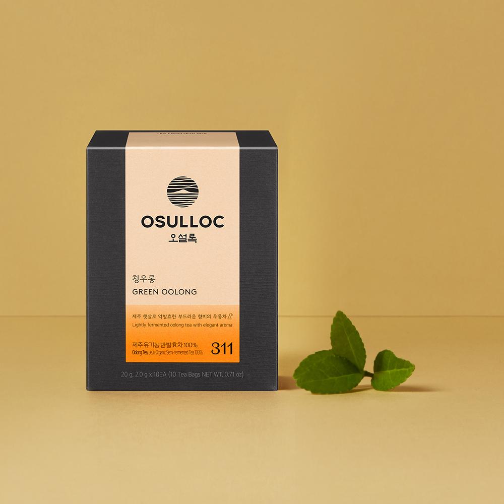 OSULLOC Green Oolong — Charcoal-grey box beside fresh green leaves on ochre.