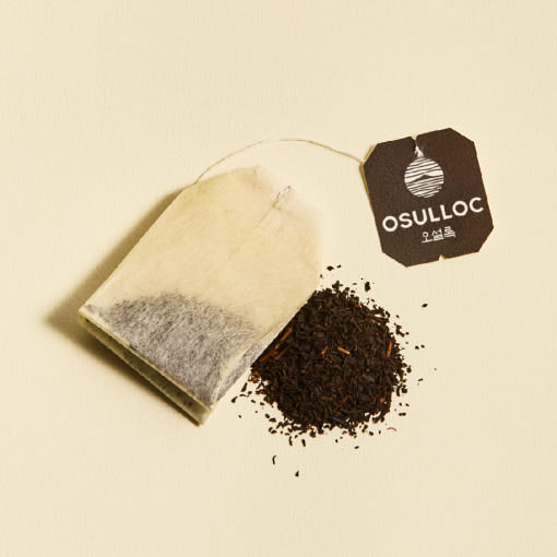 OSULLOC Yuja Earl Grey — Bolsita de papel junto a té oscuro suelto sobre amarillo.